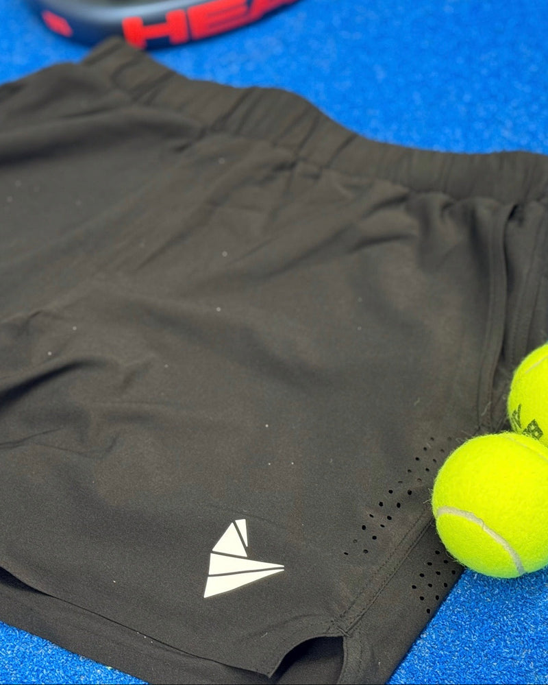 Black athletic shorts with a logo on a blue padel court with padel balls next to them.