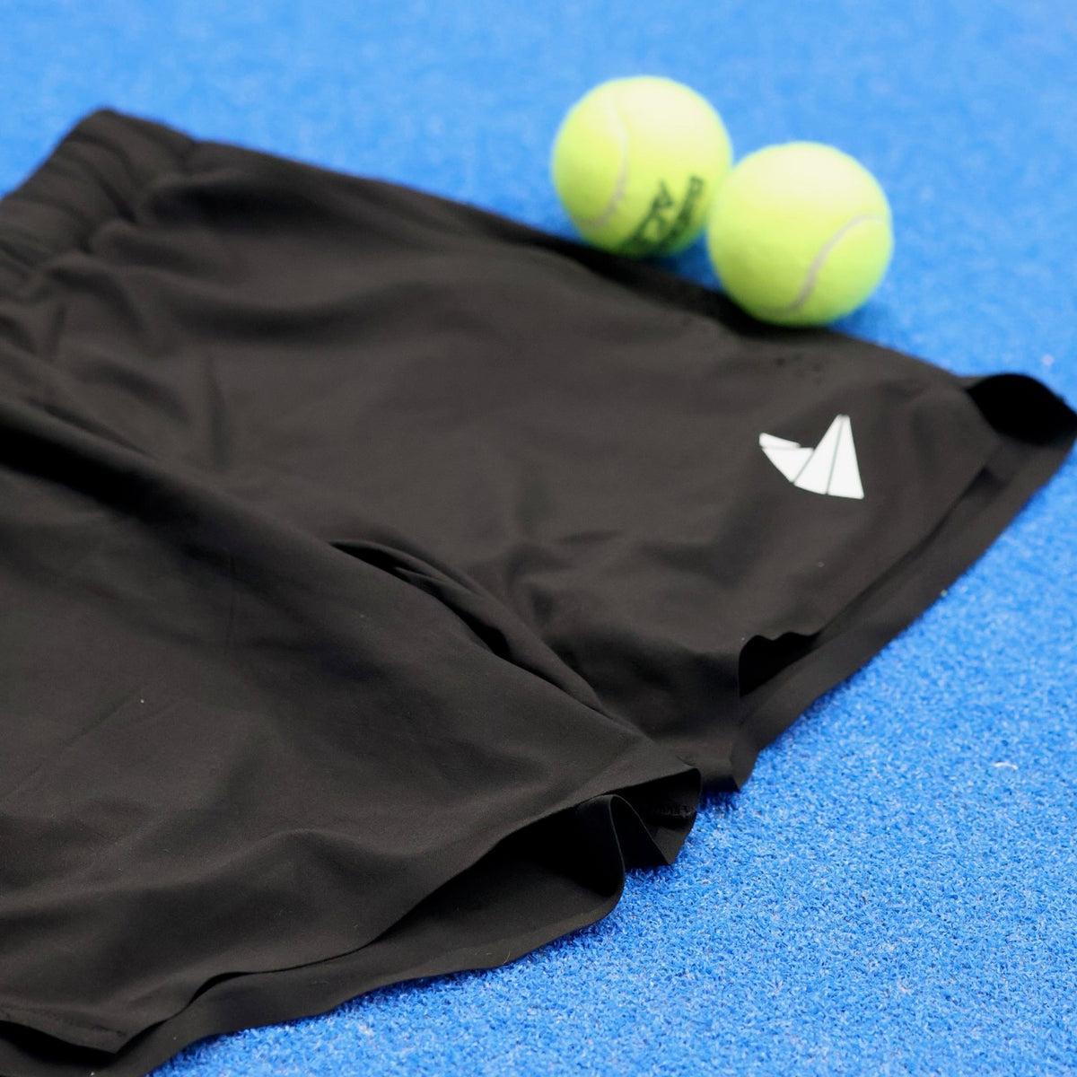 Black atheltic, comfortable, padel shorts on a blue padel court next to some padel balls.